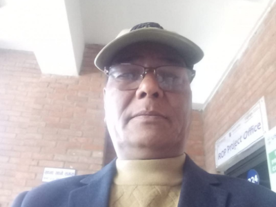 Elderly man wearing a beige cap and glasses, standing against a brick wall.